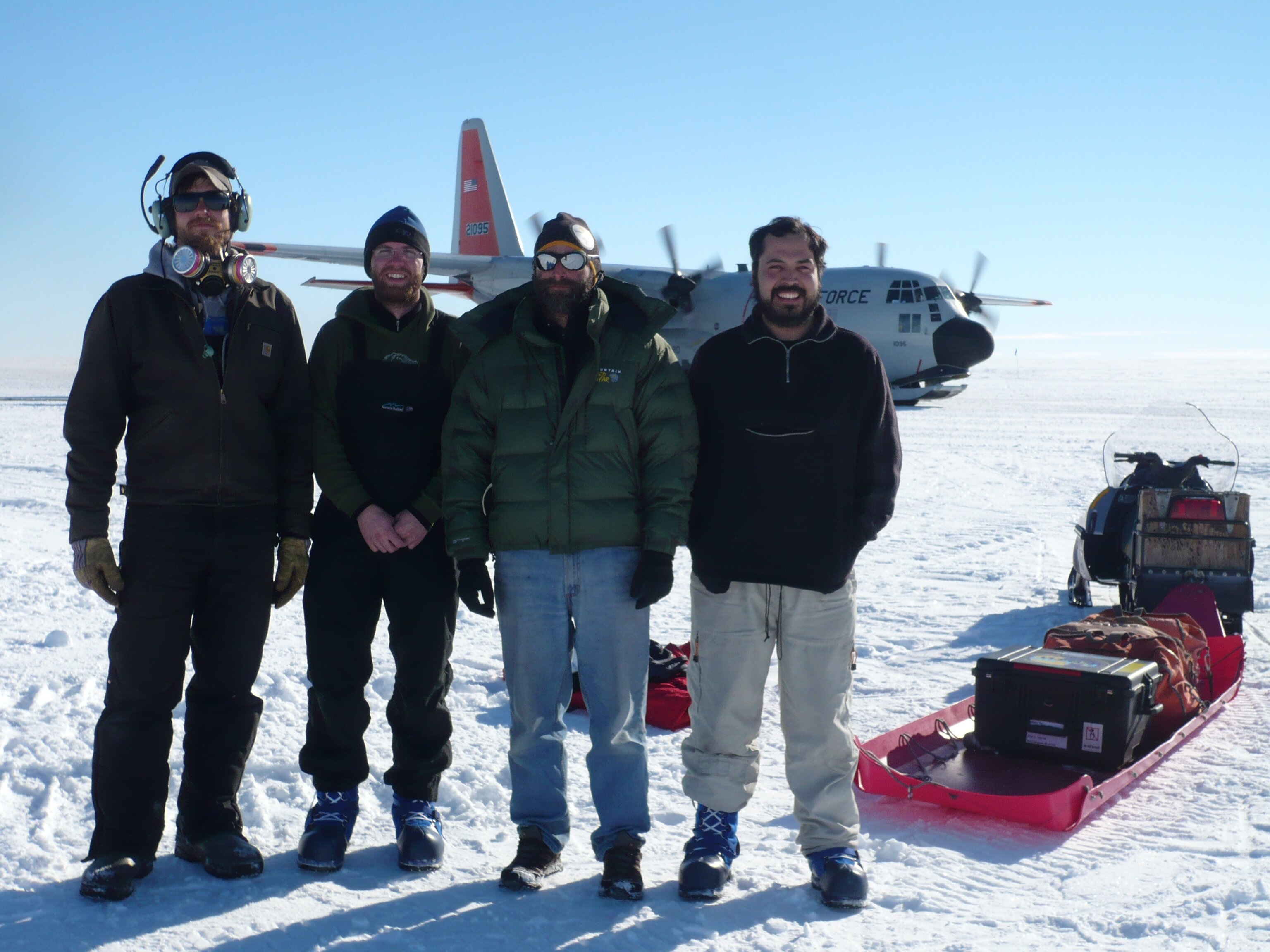 Four men stand in front of a plane and a sled with equipment in a snowy, remote location. Photo by Cameron Lewis